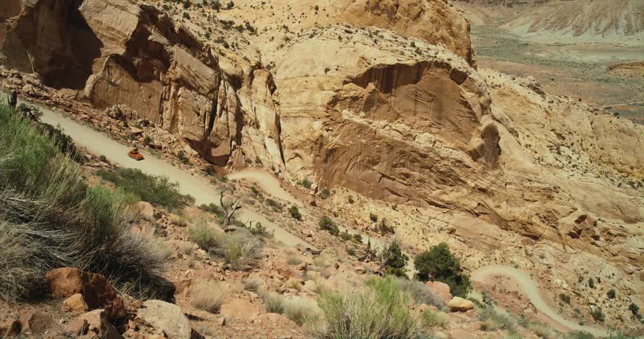 A vehicle travels along a winding dirt road down a mountain slope in an arid countryside during the day
