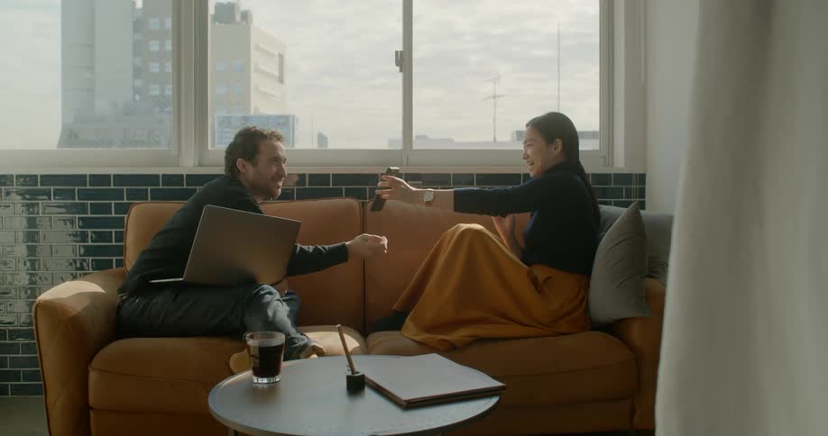 A woman sitting on a couch with a man shows him her cellphone as he looks at his laptop on his knee