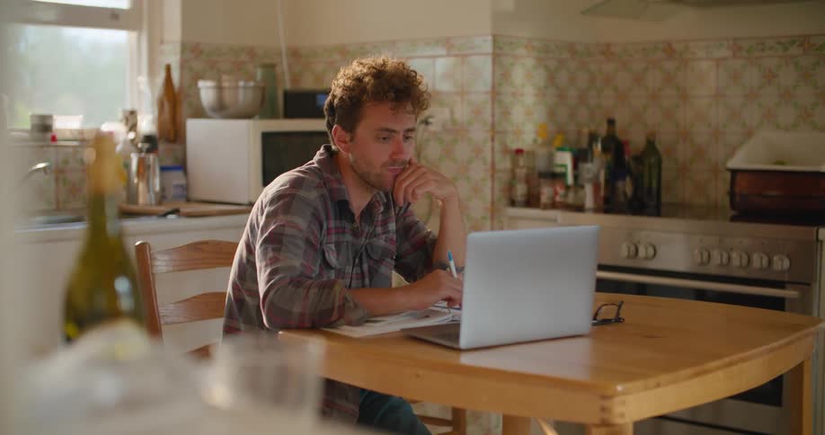 Man sits at wooden table in kitchen contemplating laptop