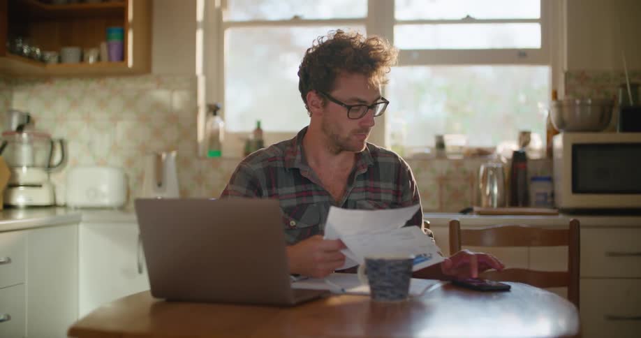 Man sits at kitchen table viewing papers and laptop screen and using calculator