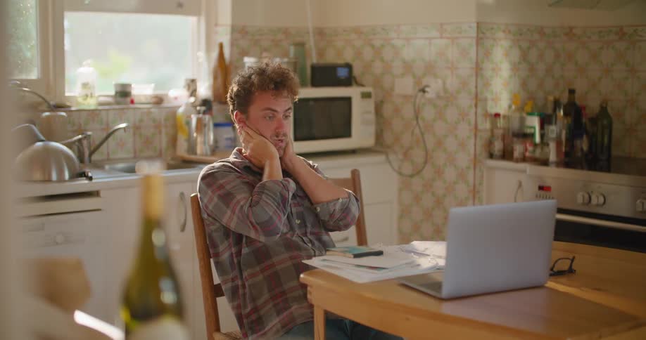 A male siting at a table near paperwork and a laptop looking stressed and worried