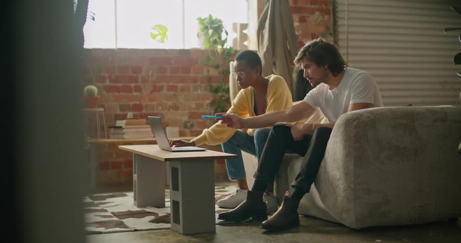 A woman operates a laptop while the man sitting next to her speaks and points at the screen with a pen while the sit together on a sofa indoors during the day