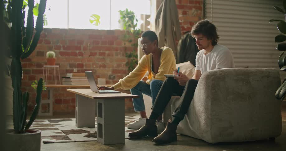 A man and a woman sitting in a room while the woman uses a laptop with a wall and window in the background