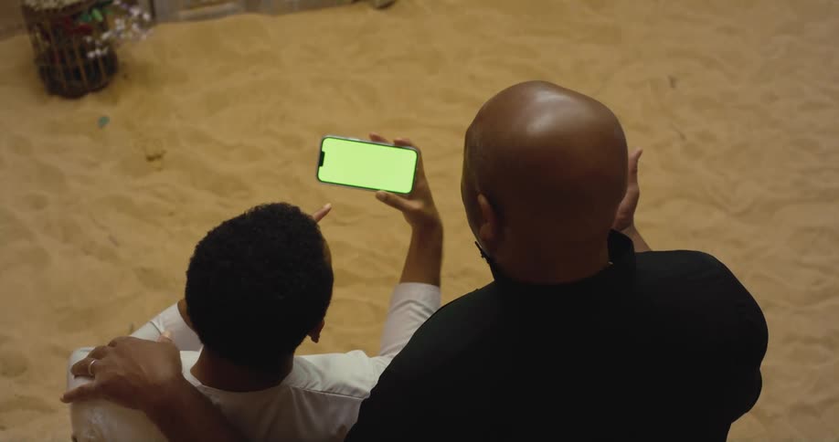 Two men situated on sand share a smartphone screen