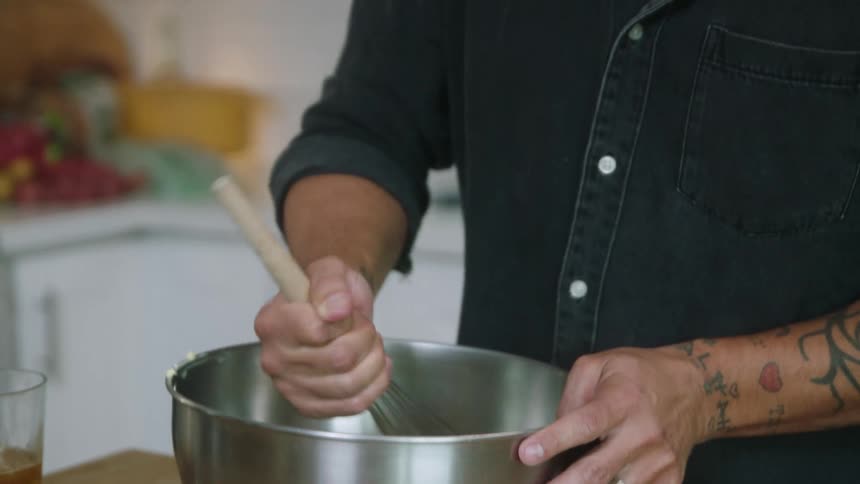 A man is stirring something in a silver mixing bowl with a wire whisk