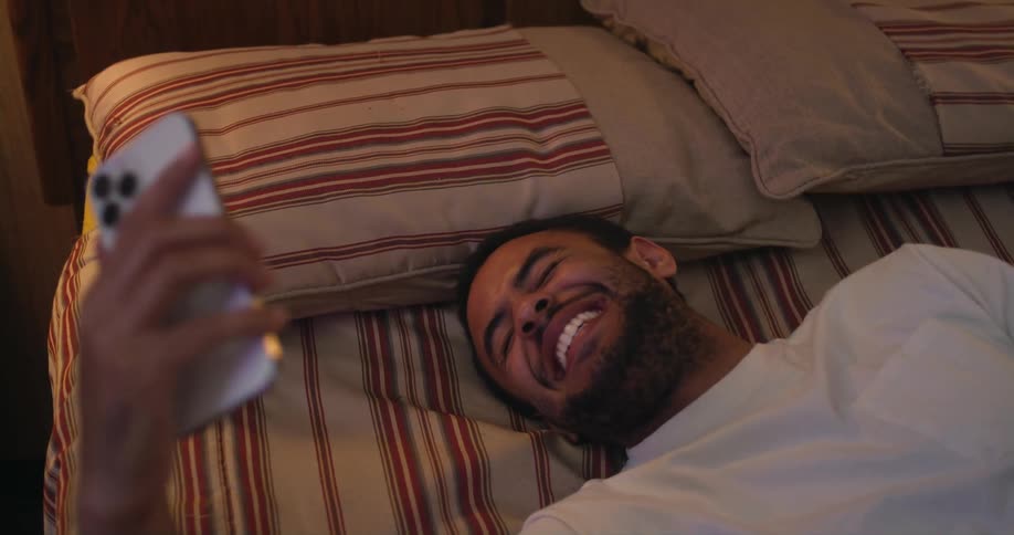 A man laughs while participating in a video chat with his smartphone as he lies in bed
