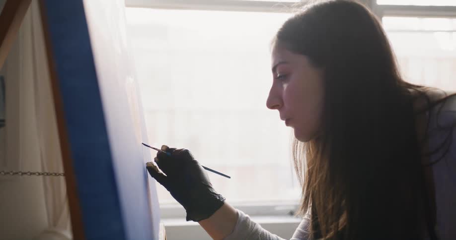 A woman wearing black gloves is standing in a room painting on an easel