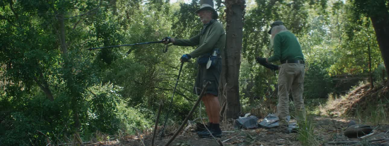 Two elderly men stand in a forest one points with his walking sticks and talks as the other shakes a net