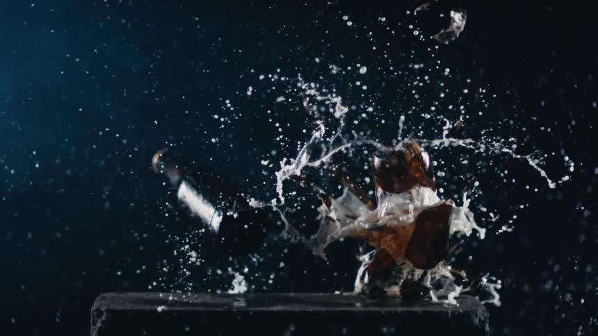 Beer bottle falling on to a stone and smashing open as beer flies in to the air