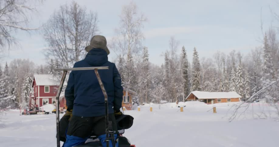 A man stands as he rides a snowmobile along deep snow in a street near some houses