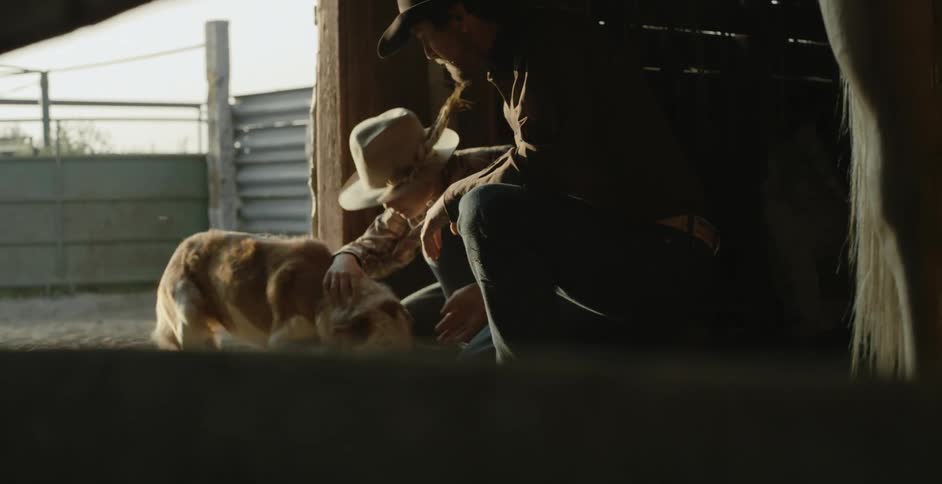 A father and son in a barn call a dog over and bend down to pet the dog
