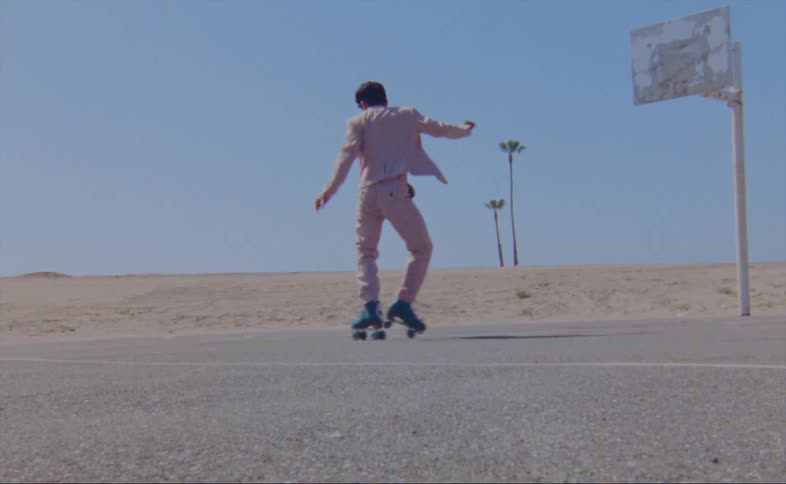 A man wearing a pink jacket and pants is dancing on roller skates on an outdoor basketball court