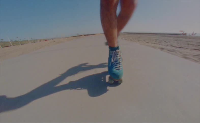 A man wearing shorts and a striped shirt is skating backwards doing cross cuts on a road at the beach