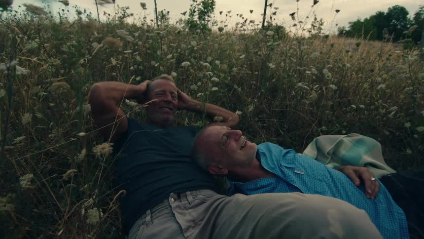 A man laughs while lying with his head on the chest of another man in a field of tall grass on a cloudy evening