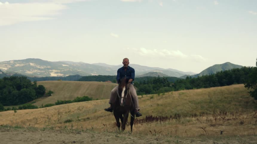 Man with blue shirt and brown pants rides brown horse uphill
