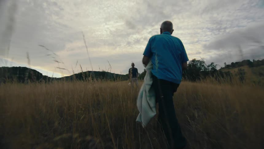 A man carries a blanket while walking through a field of tall grass towards another man waiting for him