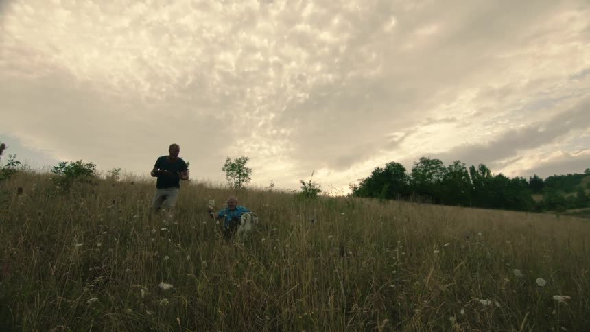 A man walking through a field alongside another man trips and falls before beginning to laugh