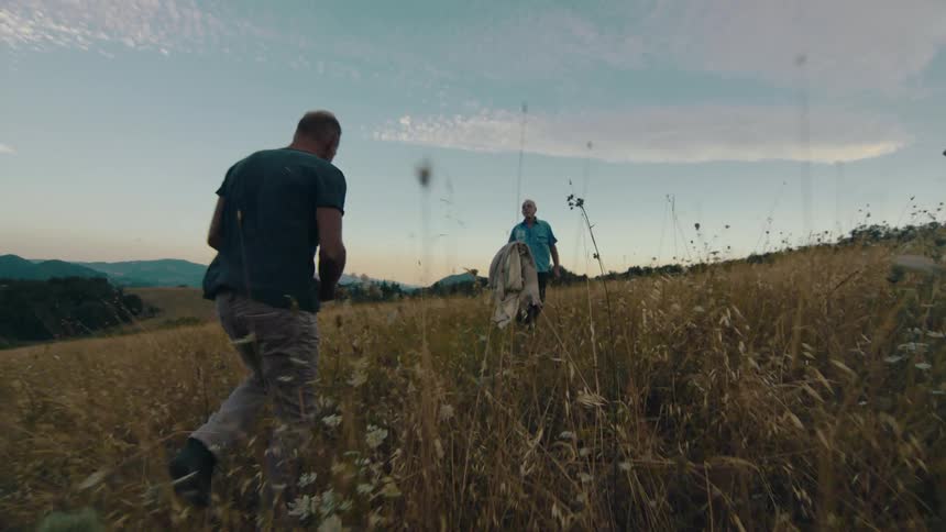 A man carrying flowers laughs while walking through a field of tall grass towards another man