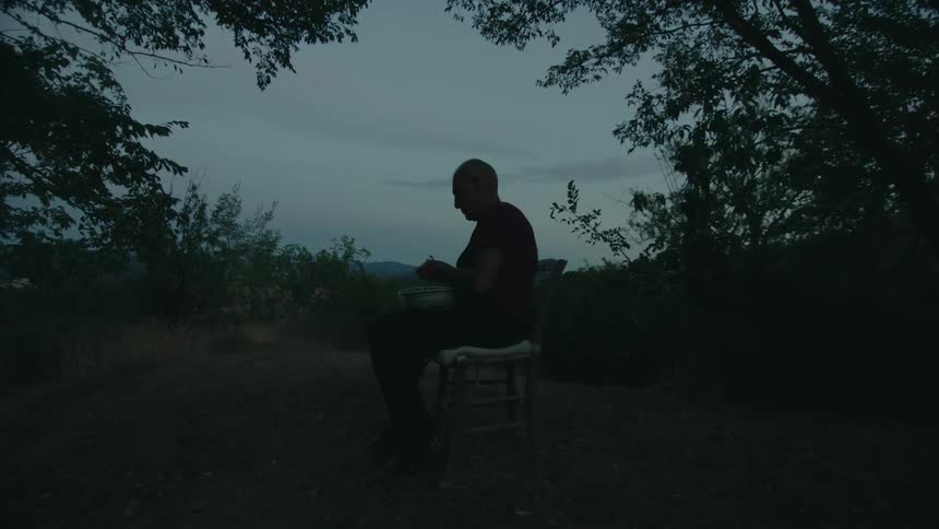 A man peels potatoes while sitting in a chair outdoors in the evening