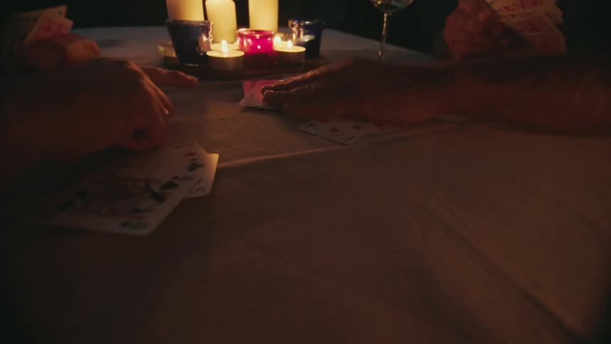 Two men play a card game at night sitting at a table lit by candles