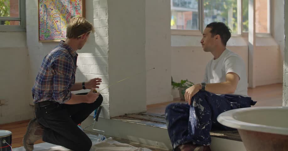 A man uses a measuring tape to measure a section of flooring while speaking to another man indoors during the day