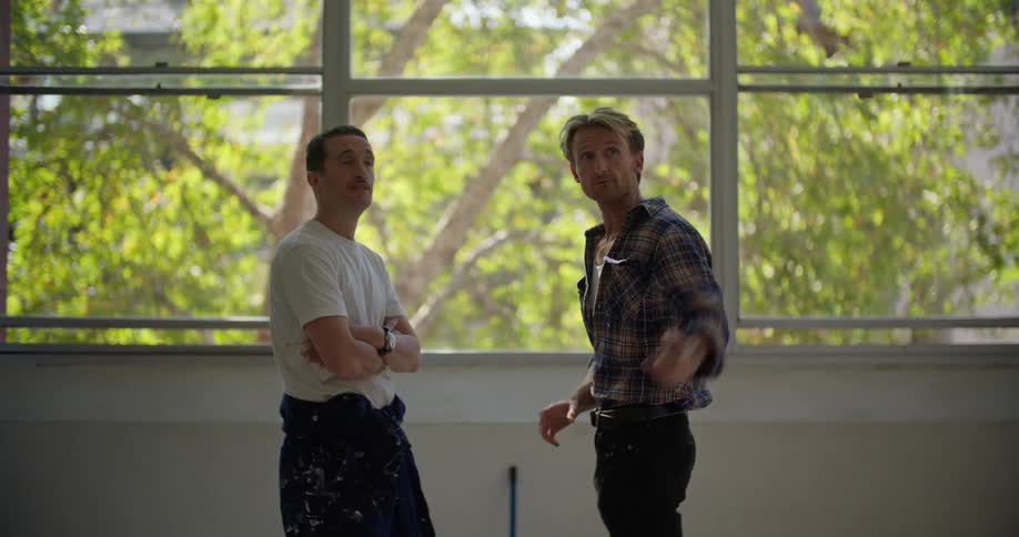 A man gestures out of frame while speaking to another man before a large window indoors during the day