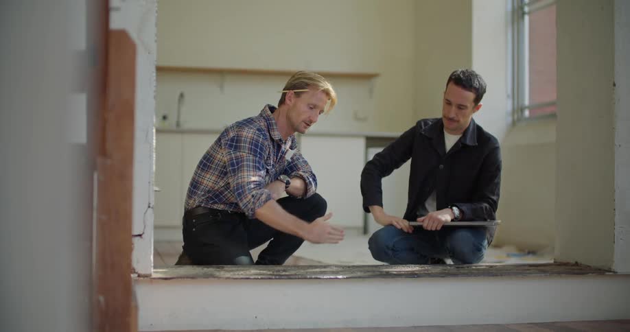 A man shakes hands with a contractor as they kneel on the floor of an apartment to discuss plans