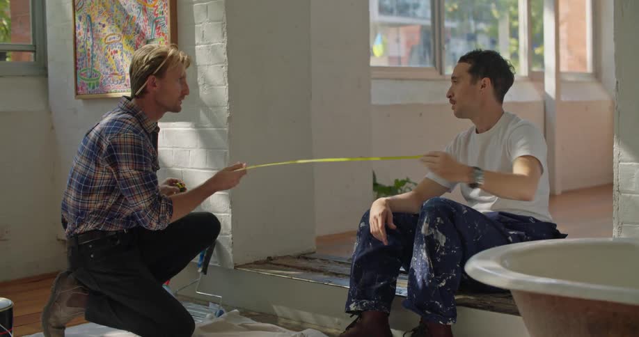 A pair of men talk while using a measuring tape to measure a wide doorway in a house during the day