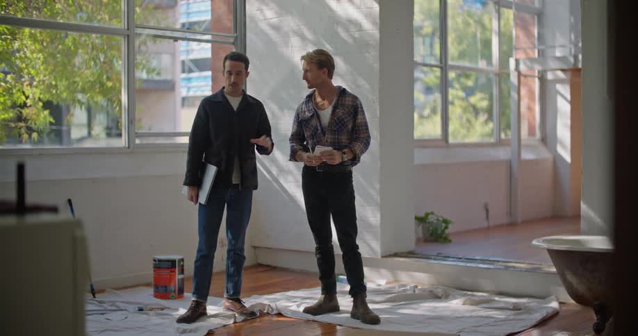 Two men are standing in a room talking and there is a drop cloth on the floor and one make has a book under his arm and the other is holding a notepad and pen