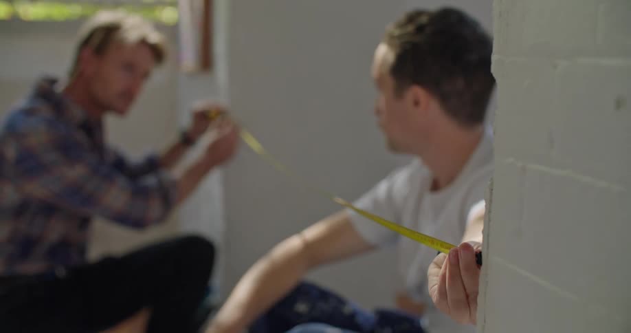 Two men are in a room talking and they each hold an end of a tape measure across a hallway