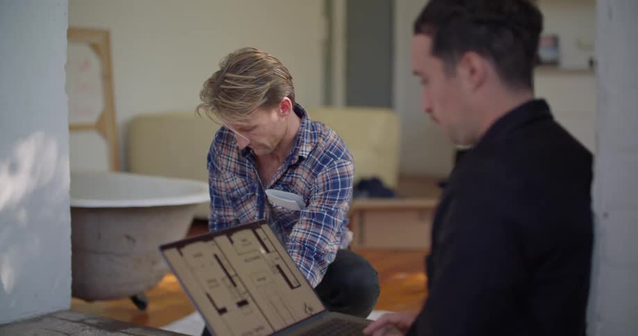 A man viewing a floorplan on a laptop talks to a man using a measuring tape to measure a section of flooring