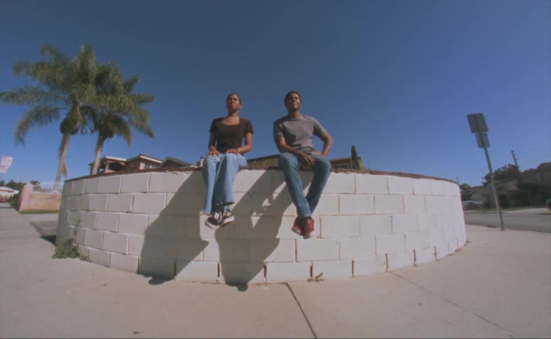 A man and a woman are sitting outside on a ledge in a neighborhood near palm trees under a clear blue sky