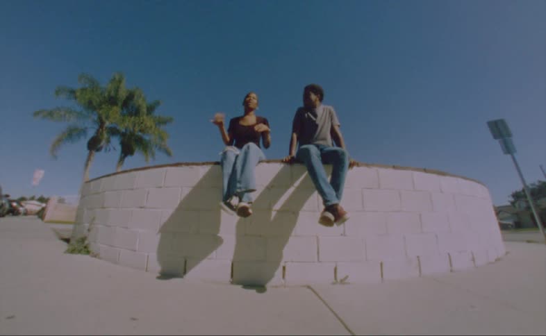 A man and a woman are sitting outside on a ledge talking in a neighborhood near a palm tree under a clear blue sky