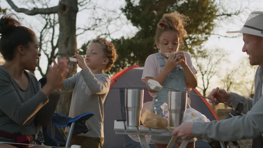 A woman claps while watching her kids make and eat smores while camping
