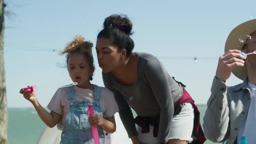 A woman and a girl bubbles from a bubble wand together while standing outdoors on the coast during the day