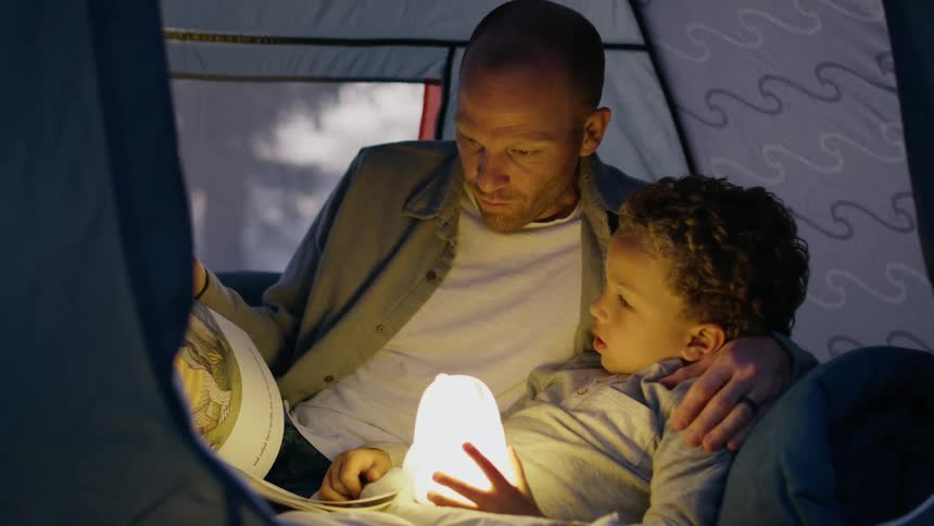 A father lies with his son in a tent and reads a book to him