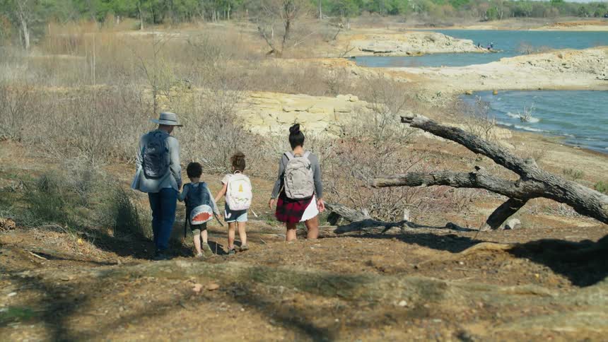 A family of four all wearing backpacks walks down a small slope heading toward the sand and water