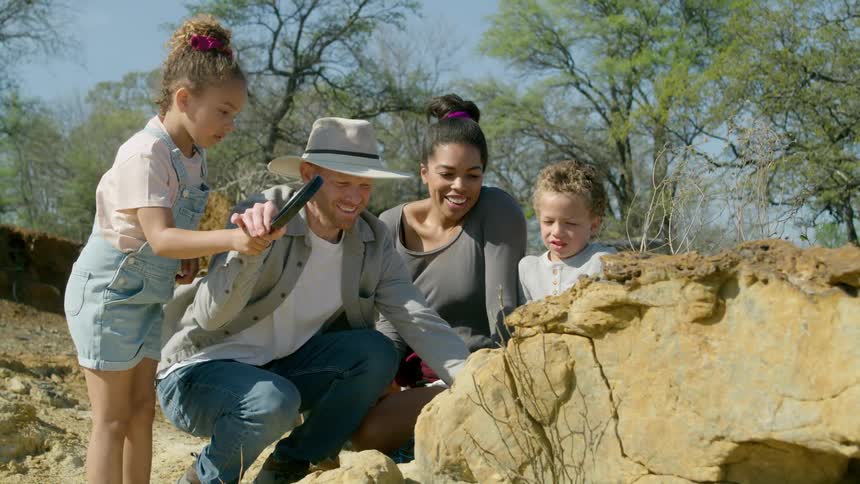 A father and daughter are holding a magnifying glass near plants and rocks as the mother and little brother watch