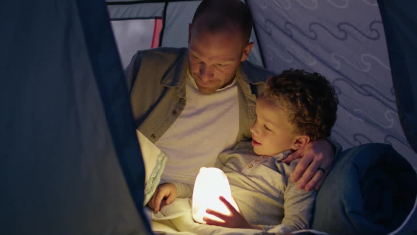 A man is in a tent with his young son and they are looking at a book together as the boy holds a light