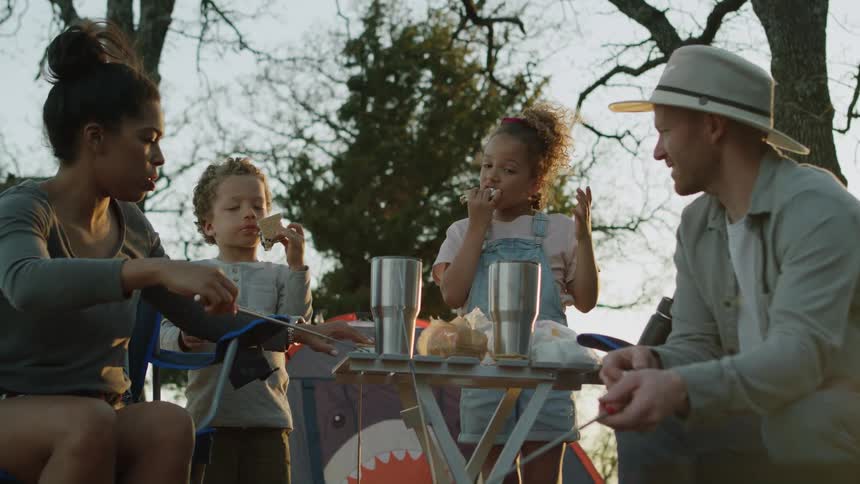 Parents cook smores on a campfire as their children eat cooked smores behind them