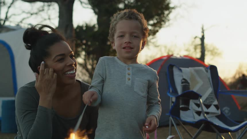 A little boy is standing outside near tents holding a sparkler and his mother is with him