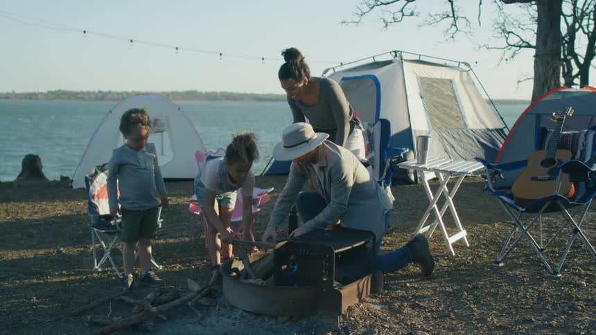 A family with two young children are camping near the water and putting firewood near the grill