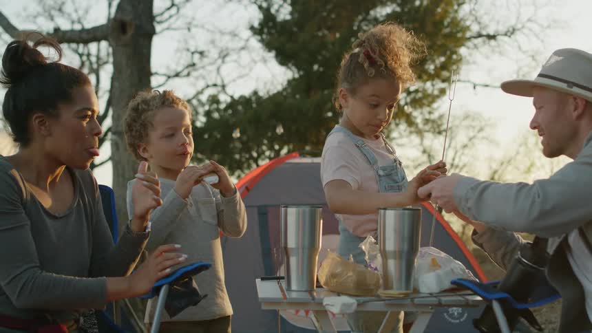 A family with two young children are camping and cooking out and eating