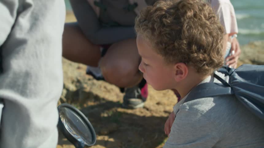 Rest of the family smiles and watches as son examines ground on shore with large magnifying glass