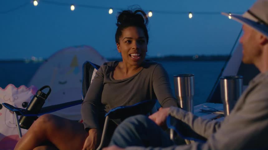 A husband and wife sit outside in front of tents and hold hands and talk in the evening