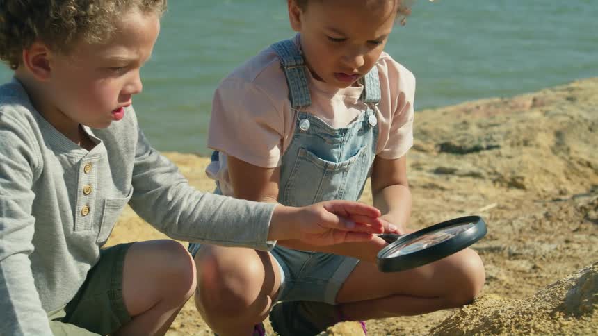 A young boy and girl sit on the shore and look through a magnifying glass at the rock
