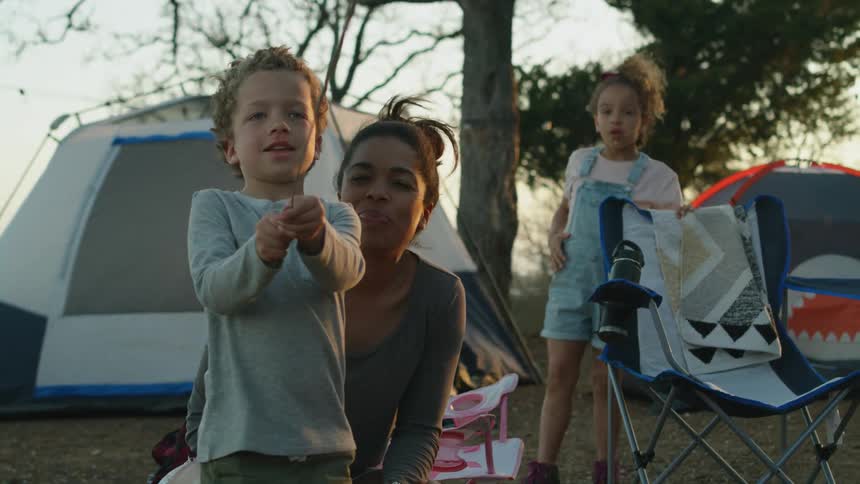 A little boy is standing outside near tents holding a sparkler and his mother and sister are behind him