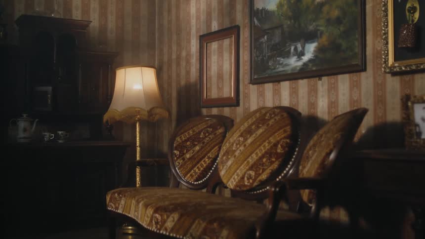 A light is on in a living room with antique furniture and an empty picture frame next to paintings on the wall