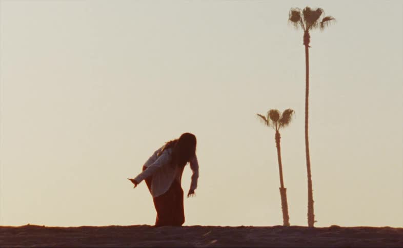 A woman wearing red baggy pants and a white shirt is outside dancing near palm trees