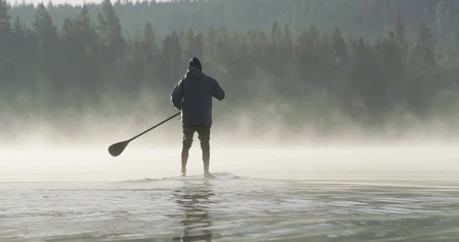 A man is standing on a board rowing over a lake